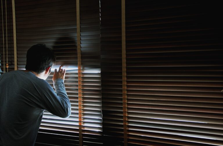 Man looking through wooden blinds from a dark room